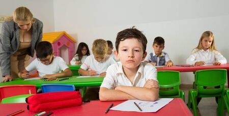 Portrait of upset boy in schoolroom on background with pupils studying with teacherの写真素材