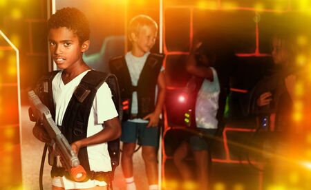 Confident tween mulatto boy standing with laser pistol in dark lasertag room during game with friendsの写真素材