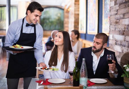 Diligent friendly pleasant waiter with dishes serving man and woman friendly company indoorsの写真素材