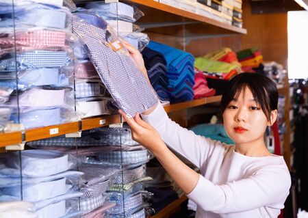 Young Chinese glad positive smiling girl looking for fashionable mens shirt in clothing storeの写真素材