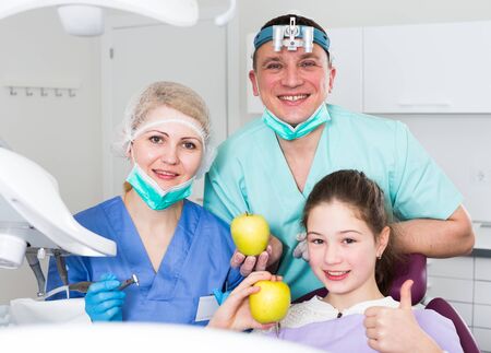 Smiling dentists standing near satisfied teen girl showing thumbs up after dental treatment, holding applesの写真素材