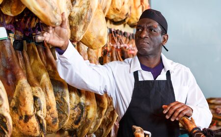 Confident African American salesman checking quality of a Spanish ham at a meat storeの写真素材