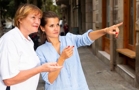 Polite young woman pointing way to middle aged female tourist on summer city streetの写真素材