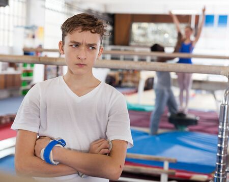 Unhappy tired teenage boy in sportswear standing near parallel bars at gymnastic hallの写真素材