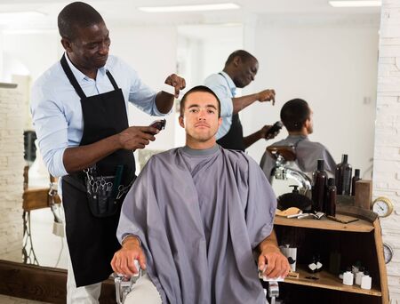 Young man getting haircutting with electric clipper by professional African barber in salonの写真素材