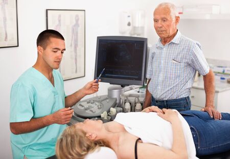 Professional efficient positive friendly sonographer explaining results of ultrasound diagnostic to female patient and her husbandの写真素材