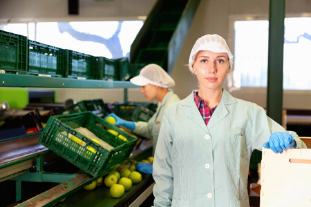 Young glad cheerful positive smiling woman in uniform during sorting at warehouse at apples factory