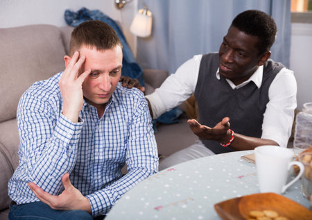Offended adult man sitting at home table while his African American friend soothing him