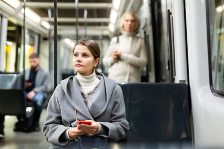 Portrait of positive young woman using mobile phone while traveling by public transport in autumn...