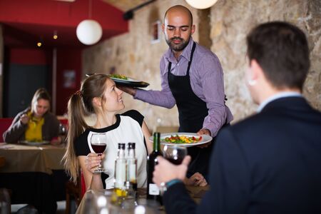 Polite waiter bringing ordered dishes to young smiling couple at restaurantの写真素材