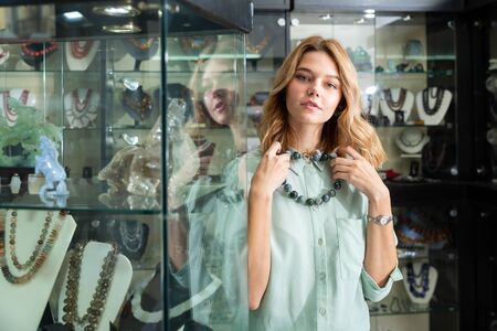 Young smiling female client choosing natural gemstone jewelry in boutique, trying on beads made of grey agateの写真素材