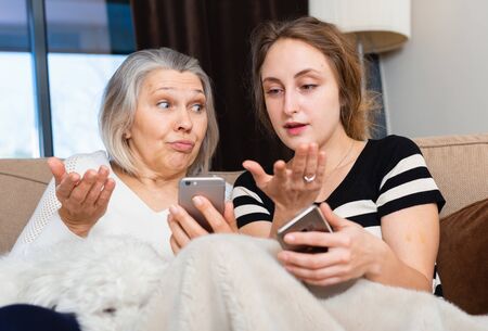 Mature woman and adult daughter using phones sitting on sofa at homeの写真素材