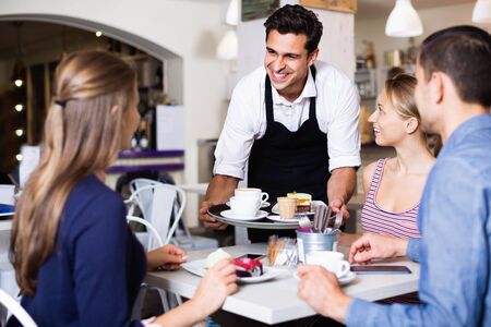 Smiling diligent waiter showing hospitality and serving visitors in cozy coffeehouseの写真素材
