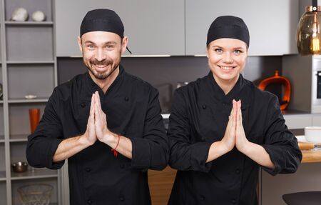 Portrait of two smiling personal chefs in black uniform greeting in thai style in kitchen interiorの写真素材