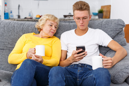 Concentrated young guy using smartphone and his mother sitting near him