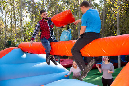 Guys having funny wrestling by pillows on inflatable beam in outdoor amusement playground
