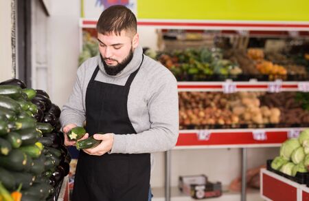 Male shop assistant lays fresh cucumber on counter in grocery shopの写真素材