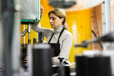 Skilled woman engaged in olive oil production, controlling process of bottling of finished product in glass bottlesの写真素材