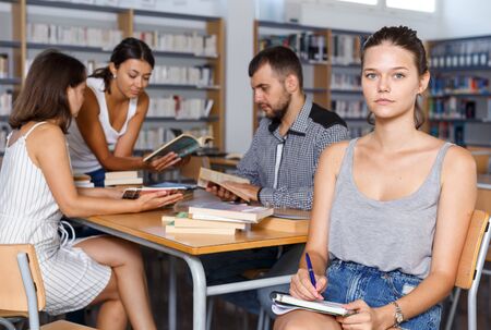 Serious female student sitting with notebook in high school library on background with working coursematesの写真素材