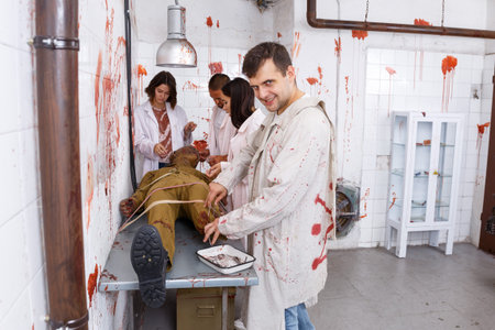 Guy with mad look standing over zombie mannequin with friends in quest room styled as abandoned...