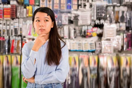 Portrait of positive glad Asian girl having doubts about choice of goods in cosmetics storeの写真素材