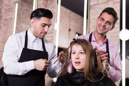 Attractive long-haired brunette getting haircutting from two professional hairstylists in salonの写真素材