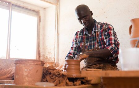 Portrait of positive afro man making ceramic pot on pottery wheelの写真素材