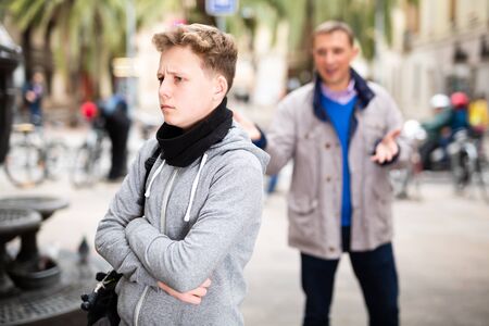 Frowning teenager listening to reprimanding from his father on city streetの写真素材