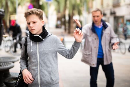 Frowning teenager gesturing enough while adult man reprimanding him on city streetの写真素材