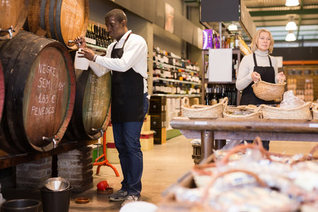 African American seller pouring draft wine from wooden barrel to plastic can while mature saleswoman arranging showcase with bulk food in grocery shopの写真素材