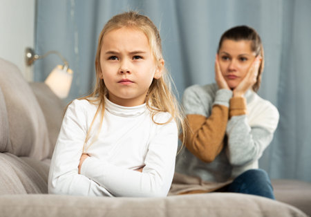 Frustrated small girl sitting at sofa,  having conflict with mother at home interior