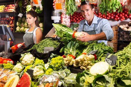 Portrait of young positive male in apron selling fruits and vegetables in shopの写真素材