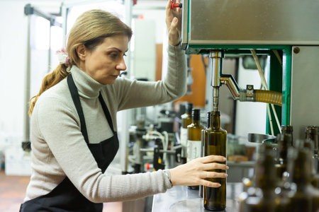 Skilled woman engaged in olive oil production, controlling process of bottling of finished product...
