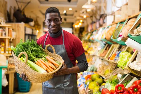 Positive African American man owner of greengrocery shop in apron offering fresh carrots for saleの写真素材