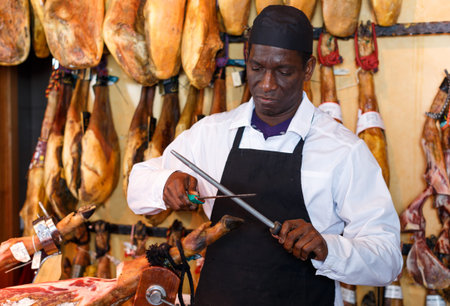 Confident African American salesman sharpening knife for cutting delicious ham in butcher shop