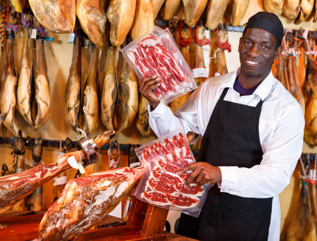 Smiling African American seller offering a delectable spanish jamon in butcher shop