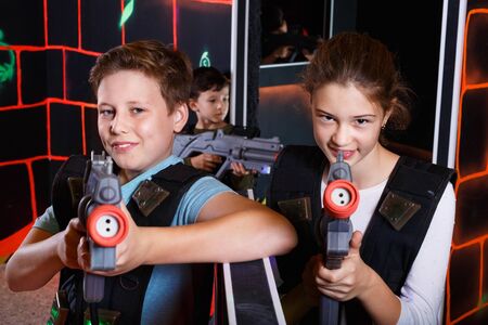 Cheerful happy  teen girl and boy with laser pistols playing laser tag with friends on dark labyrinthの写真素材