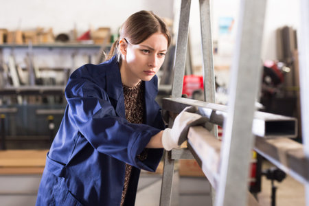 Confident young woman in blue overalls working in mechanical metalworking workshop, checking metal...