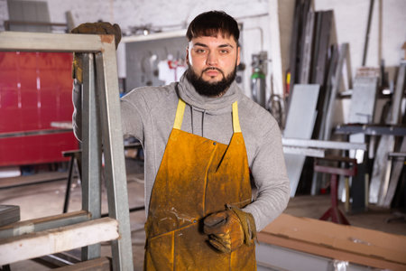 Serious young bearded locksmith posing in interior of small private workshop