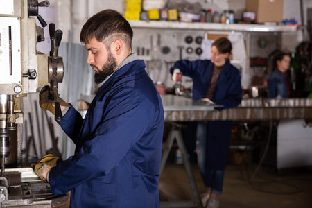 Serious young craftsman working on metal structures drilling machine in industrial workshop