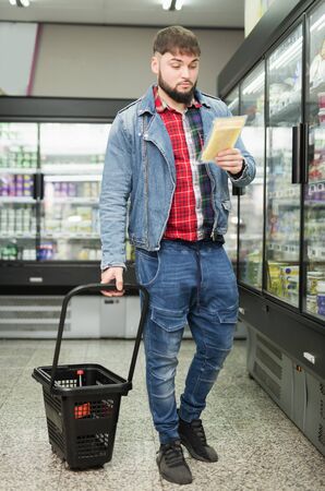 Interested cheerful young man shopping in grocery store, looking for hard cheeseの写真素材