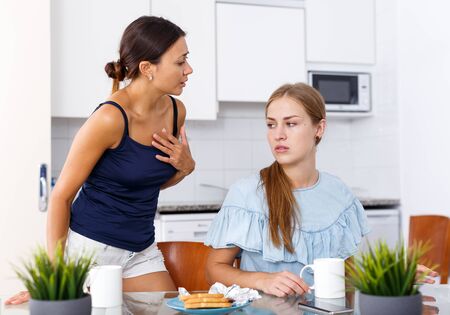 Young woman and her sad friend finding out relationship after quarrel at kitchenの写真素材