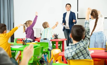 Smiling young woman teacher examining pupils at lesson in elementary school. Children raising hands to answerの写真素材