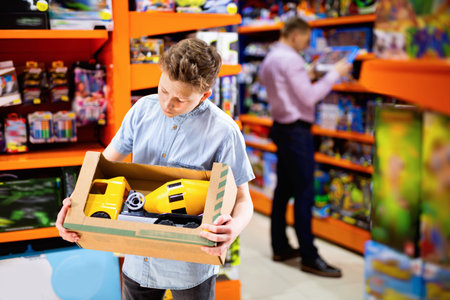 Serious happy cheerful positive smiling teen boy looking narrowly at yellow plastic car in children toy store