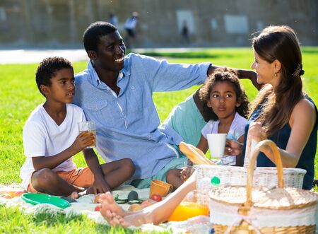 Portrait of cheerful interracial family with two children enjoying picnic on green meadowの写真素材