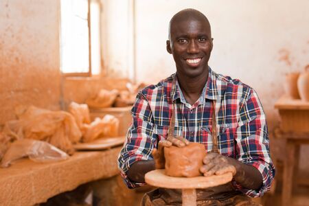 Smiling African American guy enjoying work with clay on potter wheel in ceramics workshopの写真素材