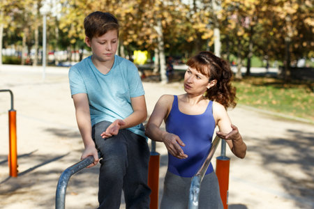 Athletic family of mother and preteen boy training together outdoors, doing exercises on parallel...