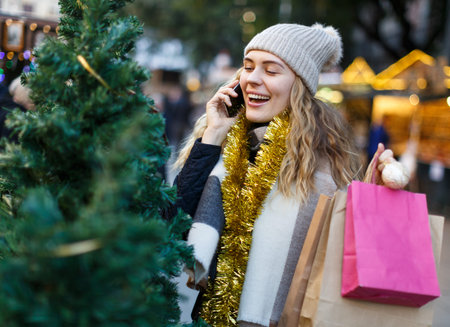 Smiling young woman spending time at Christmas fair and talking on mobile phone