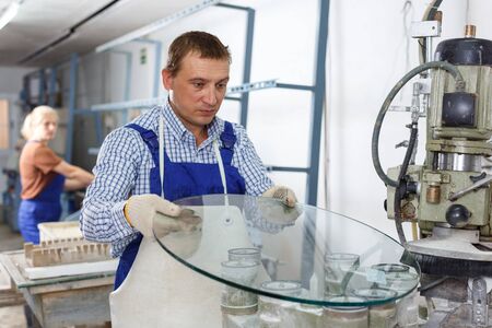 Experienced male worker of glass factory engaged in processing of curved glass beveling on industrial machineの写真素材