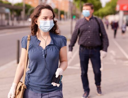 Focused woman wearing medical mask and rubber gloves listening to music on earphones on way to work along city street on spring day. New life reality during COVID 19 pandemicの写真素材